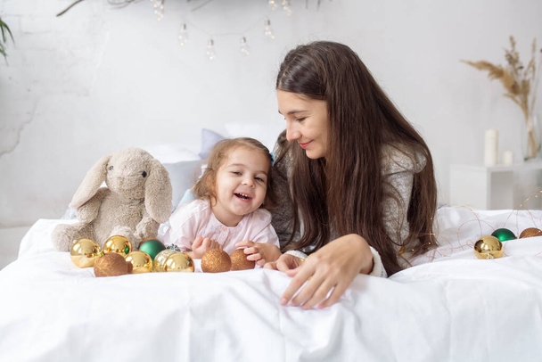 A beautiful curly girl 2 years old with her young mother lie on the bed among garlands and decorations and laugh - Foto, Imagen