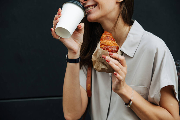 Smilng mature woman on the street drinking coffee with wrapped in paper croissant. Close up, cropped face. - Photo, Image