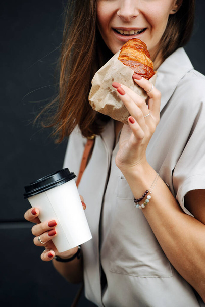 Smilng mature woman on the street eating wrapped in paper croissant with cup coffee. Close up, cropped face. - Фото, зображення