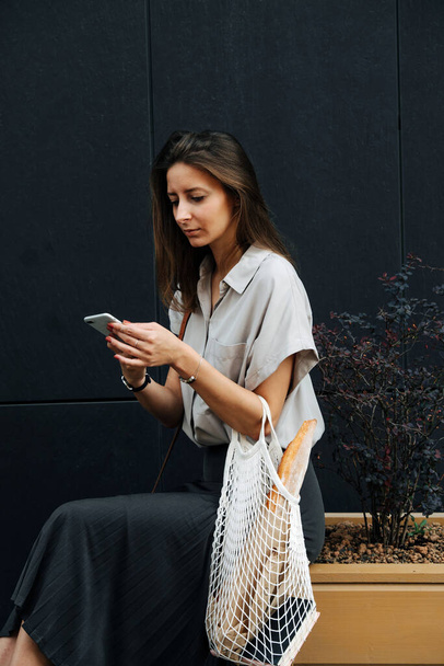Mature woman in corporate clothing standing on the street. She's looking at her phone, sitting on a planter box with bonsai tree, holding net bag with baguette. - Foto, Imagen