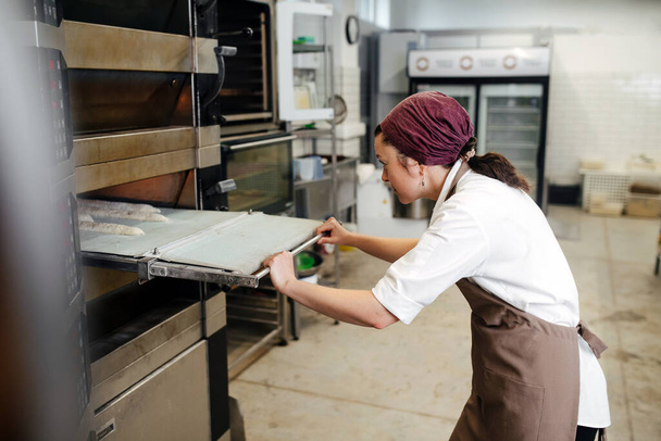 Keen female baker placing flowered baguettes in oven furnace in a bakery. Side view. - Foto, afbeelding