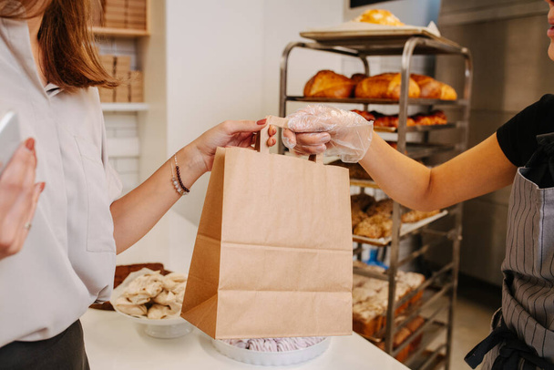 Woman making a purchaise, receiving paper bag from a baker standing behind cash register. Cropped, no heads. - Fotoğraf, Görsel