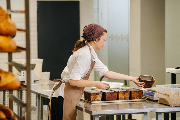Happy focused woman working in a bakery, placing bread forms on a metal tray. Side view. - Foto, Bild