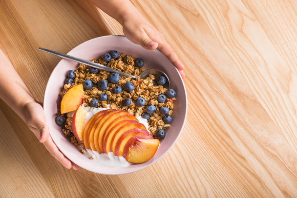 Schaal granola met yoghurt, perzikschijfjes en bosbessen op een houten tafel. kind dat 's ochtends ontbijt. - Foto, afbeelding