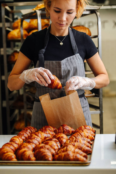 Baker woman placing freshly baked crispy golden croissants in a paper bag for delivery. Frontal view - Fotografie, Obrázek