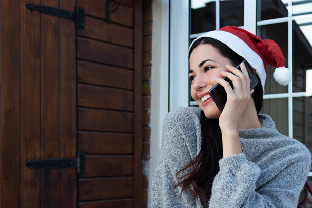 Een jonge vrouw belt met haar mobiele telefoon terwijl ze glimlacht met een kerstman hoed op Kerstmis - Foto, afbeelding
