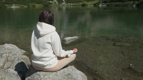 Da la vuelta a la joven en una sudadera blanca practicando yoga en una orilla del lago con agua transparente con una vista increíble de la cascada con espacio para copiar. Meditación y recreación en la naturaleza entre - Metraje, vídeo