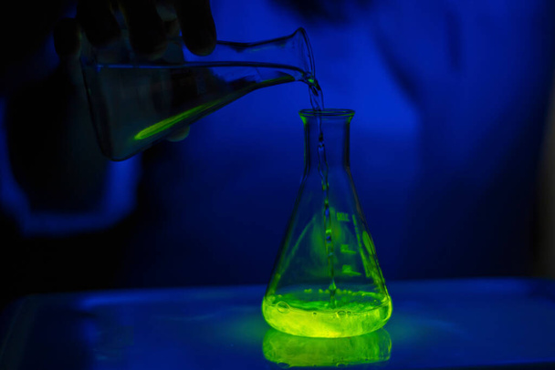 Tiedemies, joka kokeilee vihreää fluoresoivaa materiaalireaktiota erlenmeyerpullossa pimeän biokemian laboratoriossa terveydenhuollon kehittämiseksi - Valokuva, kuva