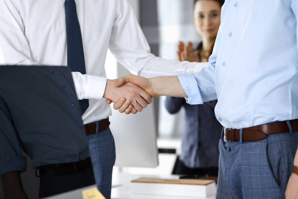 Two businessmen are shaking hands in office, close-up. Happy and excited business woman stands with raising hands at the background. Business people concept - Photo, image