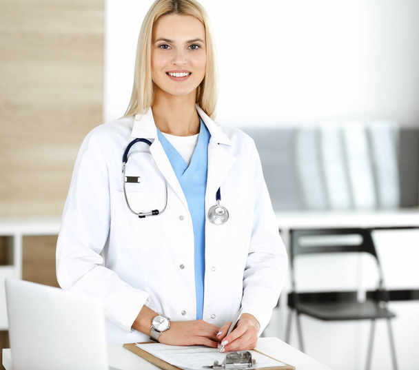 Woman-doctor at work in clinic happy of her profession. Blond female physician controls medication history record and medical exam results. Medicine concept - Foto, Bild