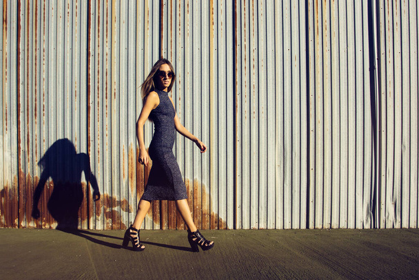 Θηλυκό νεαρό μοντέλο που περπατά με μακρύ φόρεμα σε βιομηχανική περιοχή με σκουριασμένο φράχτη στο βάθος - Φωτογραφία, εικόνα