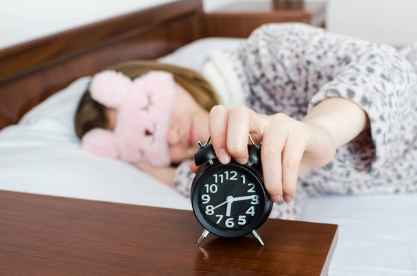 Young woman girl in cute sleep mask turns off alarm clock waking up in the morning from a call. Student do not want to wake up early for univercity. Oversleep, not getting enough sleep concept. - Photo, Image