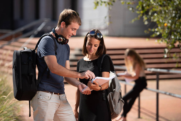 Junge lächelnde College-Studenten mit Rucksäcken arbeiten draußen auf dem Campus an einem Notizbuch. - Foto, Bild