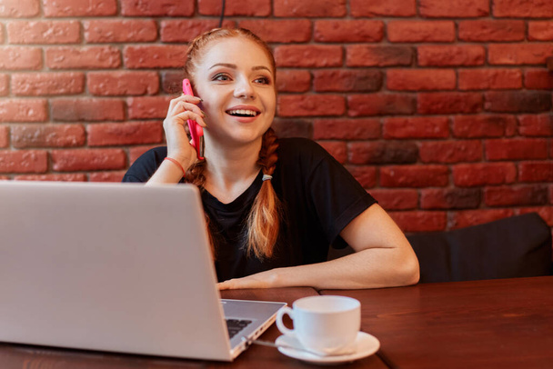 Young woman talking on phone while working online via laptop in cafe, sitting at table and drinking coffee, posing against brick wall, wearing black casual t shirt. - Photo, image