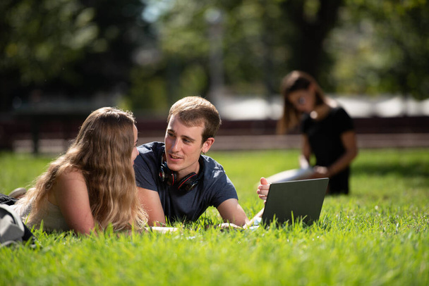 キャンパス公園の地面に寝そべっておしゃべりしている大学生のカップル. - 写真・画像