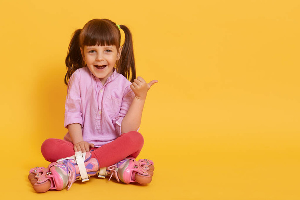 Happy girl in roller skating sitting on floor against yellow background showing thumbs aside with excited facial expression, copy space for advertisement or promotion. - Fotó, kép