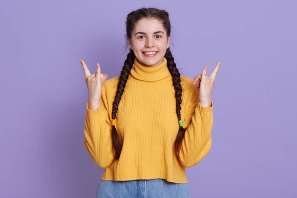 Young brunette woman posing isolated over purple background, showing rock gesture with fingers and smiles happily, wearing yellow sweater, expressing positive emotions. - Valokuva, kuva