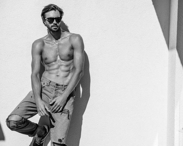 Portrait of sporty handsome strong man. Healthy athletic fitness model posing near white wall in jeans. Confident sexy fashion male with naked nude torso. Lambersexual outdoors in sunglasses - 写真・画像