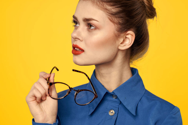 Portrait d'une femme avec des lunettes dans les mains sur un fond jaune et dans une chemise bleue vue recadrée - Photo, image