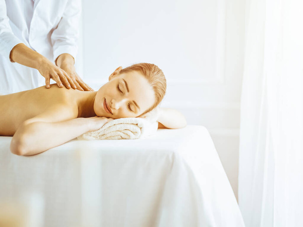 Beautiful woman enjoying back massage with closed eyes in sunny spa center - Photo, Image
