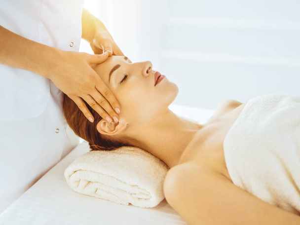 Beautiful woman enjoying facial massage with closed eyes in sunny spa center. Relaxing treatment concept in medicine - Foto, Imagem