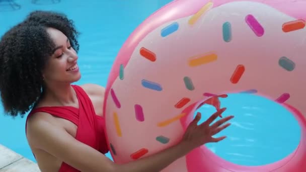 Close up view of playful African model play throw up inflatable donut in mid-air laughing looking at camera. American girl sitting on poolside toss up and spin a rubber ring smile and feel carefree - Footage, Video