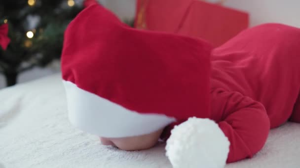 merry xmas and happy new year, infants, childhood, holidays concept - close-up 6 month old newborn baby in santa claus hat and red bodysuit on his tummy crawls with decorations balls on christmas tree - Кадри, відео