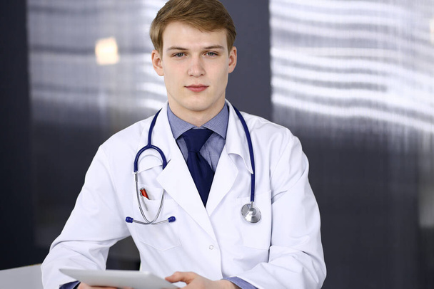Blond male doctor sitting and working with tablet computer in clinic at his working place, close-up. Young physician at work. Perfect medical service, medicine concept - Φωτογραφία, εικόνα