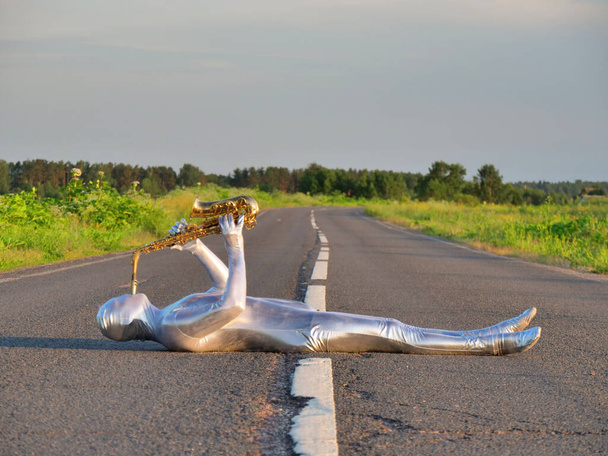 Muž hudebník saxofonista v plné tělo objímající stříbrný a stříbrný elektrický oblek drží zlatý alt saxofon, leží na prázdné silnici v létě s perspektivou. Zrůda, neobvyklá osoba, mimozemšťan, UFO. - Fotografie, Obrázek