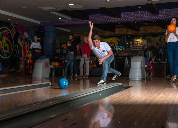 Um homem em uma pista de bowling joga uma bola azul europeia em jeans e uma camiseta branca. RÚSSIA, MOSCOW - 20 de setembro de 2020 - Foto, Imagem