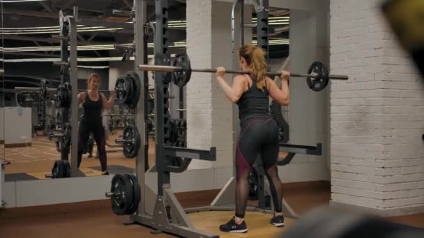 Vista trasera de sexy mujer caucásica entrenador haciendo sentadillas de barra interior de gimnasio - Metraje, vídeo