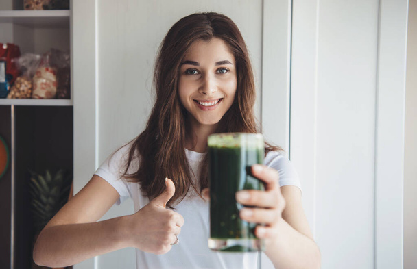 Caucasian woman holding a glass with fresh green juice is smiling and gesturing like sign at camera - Φωτογραφία, εικόνα
