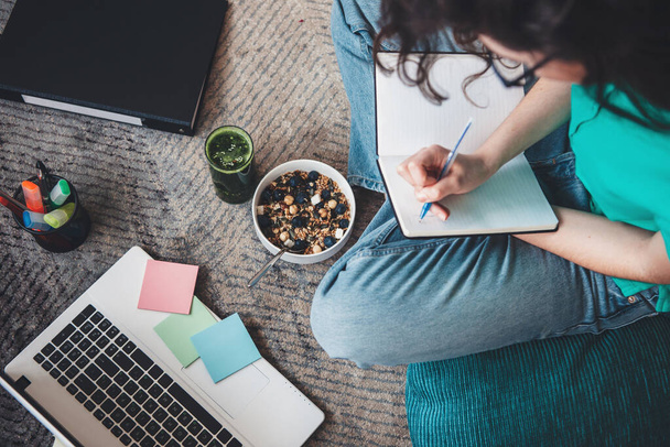 Ylempi näkymä kuva valkoihoinen nainen syö viljaa, kun kirjoitat jotain kirjaan ja käytät tietokonetta lattialla - Valokuva, kuva