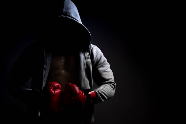 misteriosa pessoa com um capuz e luvas de boxe vermelho em um fundo escuro - Foto, Imagem