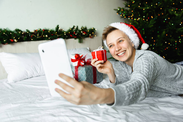 Christmas activity indoor: new year's online greetings and meetings. Women in red santa hat hold box with gift and look at a phone and communicate with friends. Anticipation of the holiday. - Φωτογραφία, εικόνα