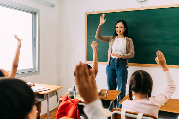 Asian school teacher with students raising hands. Young woman working in school with arm raised, school children putting their hands up to answer question, enthusiasm, eager, enjoyment - Photo, Image