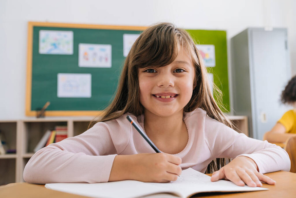 Portrait of happy female student sitting at desk in classroom smiling. Cute Caucasian elementary school girl sitting in school writing on paper, looking at camera, education, learning, happiness - Fotó, kép