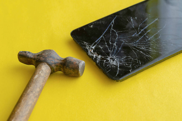 黄色の背景に壊れた黒いスマートフォンと小さなハンマー。電話やタブレットの修理サービス。選択的焦点. - 写真・画像