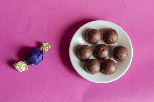 Chocolates in a white porcelain saucer on a pink background. Portion of Unwrapped Pastry and one candy in a blue wrapper. Sweet treats. Copy space. Selective focus. - Photo, Image