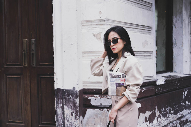 Asian pretty young hipster woman in trendy sunglasses in a vintage jacket in brown long pants with a bag stands near a white building in the city. Beautiful girl for a walk. Stylish women's clothing. - Фото, зображення