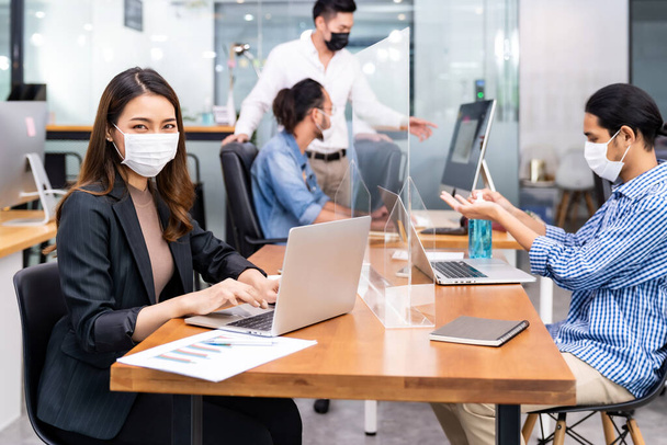 Portrait of asian office employee businesswoman wear protective face mask work in new normal office with interracial team in background as social distance practice prevent coronavirus COVID-19. - Photo, Image