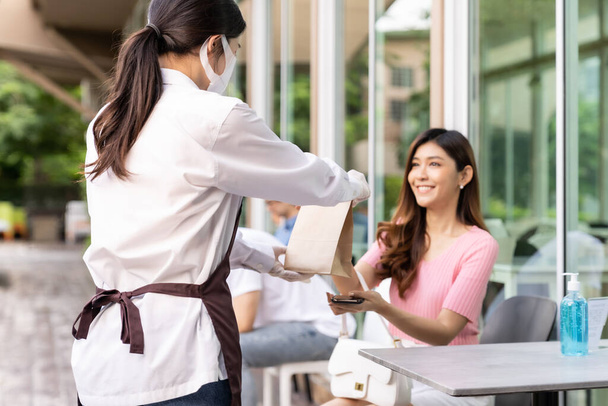 Back view of asian waitress with face mask give order of take out food bag to attractive woman female customer. Take away or take-out food service concept in new normal after coronavirus pandemic. - Фото, зображення