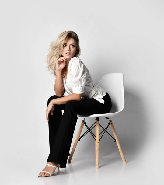 Young sexy blonde slim woman in office pantsuit shirt and pants sits on chair leaning forward and looks aside - Foto, imagen