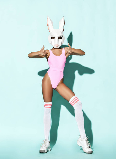 Sexy athletic slim woman model in pink swimsuit, high socks and decorative rabbit head bunny mask stands showing thumbs up - Photo, Image