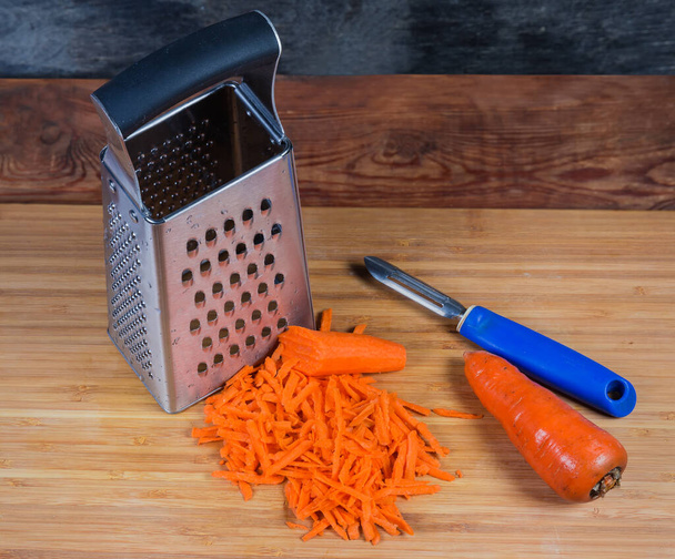 Rallador de cocina en forma de caja de acero inoxidable, pelador de verduras, zanahoria rallada en rodajas grandes y zanahoria entera sin limpiar en la tabla de cortar de madera - Foto, Imagen
