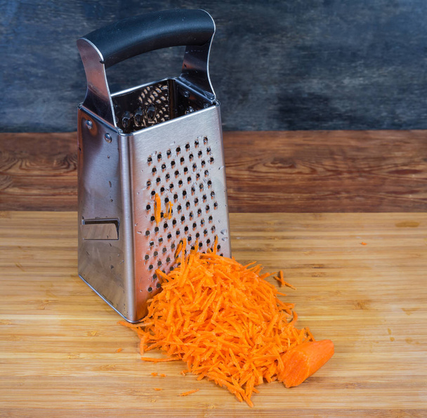Stainless steel box-shaped kitchen grater and finely grated carrots on the wooden cutting board - Valokuva, kuva