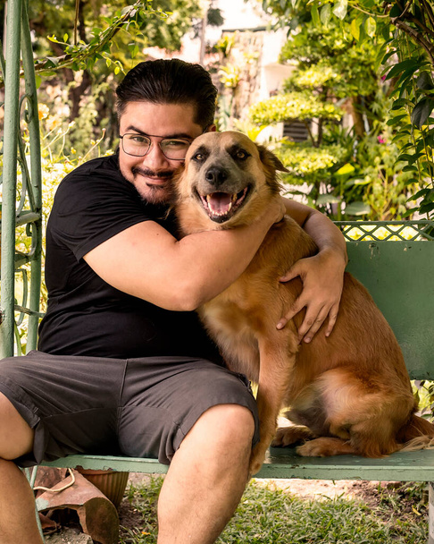 Ein bärtiger Mann im schwarzen Hemd umarmt seinen freundlichen, braunen Hund, während er an der Schaukel auf der Terrasse sitzt. Beide blicken in die Kamera. - Foto, Bild