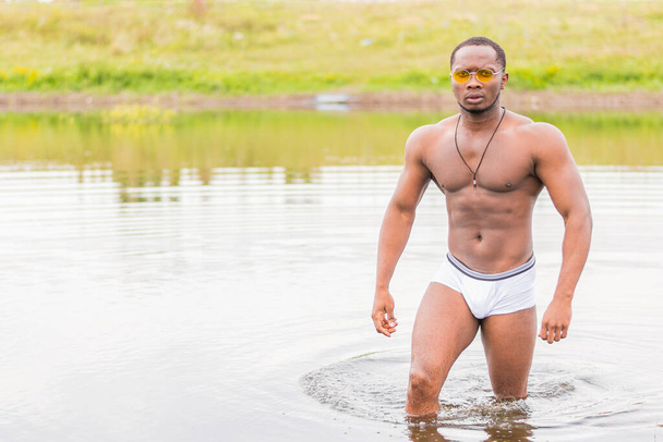 Beyaz şortlu güçlü bir genç adam sudan çıkar ve kameraya bakar. - Fotoğraf, Görsel