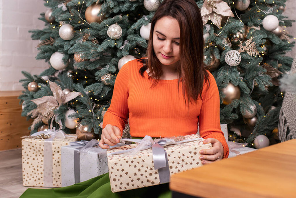 Een mooi meisje in een oranje jasje opent een nieuwjaarscadeau in een doos met tape. Kerstdag, oudejaarsavond - Foto, afbeelding