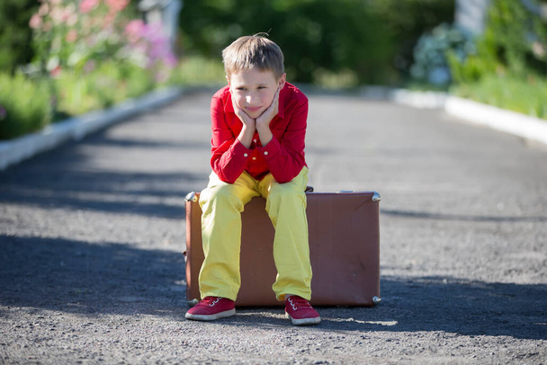 A sad boy sits on an old suitcase on the road. The child is about to leave. - Foto, imagen